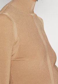 Gros plan d'une personne portant un pull à col roulé en maille côtelée couleur fauve avec des rayures texturées verticales sur les manches et le devant.