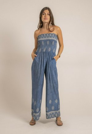 Femme portant une combinaison bleue sans bretelles avec des motifs floraux blancs, debout les mains dans les poches contre un fond clair uni.