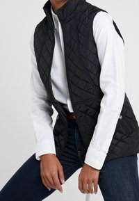 Person som bär en svart kviltad ärmlös väst över en vit skjorta med knappar och mörkblå jeans, sittande med händerna vilandes på låren.