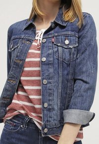 Donkerblauwe denimjack met een knoopsluiting, twee borstzakken en opgerolde mouwen. Gelaagd over een rood-wit gestreept shirt.