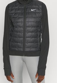 Chaleco de plumas negro con un diseño acolchado, cierre de cremallera y cuello alto. Mangas largas y negras se extienden desde la base del chaleco. Logo de Nike en el pecho.
