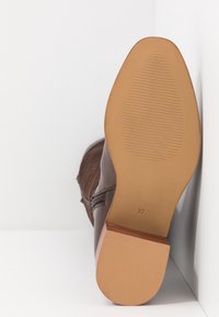 Hnědá kožená kotníková bota s hladkým povrchem, obdélníkovým podpatkem a embosovanou podrážkou. Velikost označena jako 37. Umístěna na bílém pozadí.