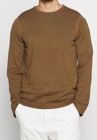 Brun långärmad tröja med ribbad struktur, rund hals och kontrasterande sömdetaljer. Bärs med vita byxor.