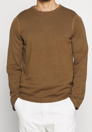 Bruine lange mouwen trui met geribbelde textuur, rond de hals en contrasterende stikdetails. Draagt met witte broek.