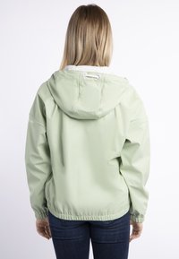 Kvinna med rakt blont hår som bär en ljusgrön huvtröja och mörkblå jeans, stående med ryggen mot en vit bakgrund.