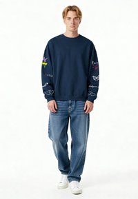 Temno modra sweatshirt s dolgimi rokavi, ki prikazuje pisane grafike na obeh rokavih. V kombinaciji z svetlo modrimi ohlapnimi kavbojkami in belimi supergami.