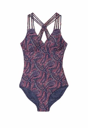 Młoda kobieta w wzorzysty jednoczęściowy strój kąpielowy biegnie wzdłuż piaszczystej plaży z spokojnym morzem i skalistym wybrzeżem w tle.