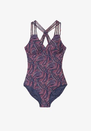 Tânără în costum de baie întreg cu model, alergând de-a lungul unei plaje cu nisip, cu mare liniștită și țărm stâncos în fundal.