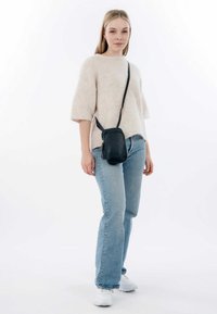 Black crossbody bag with a textured finish and tassel detail. Model wears a light beige fuzzy sweater and blue jeans, with white sneakers.