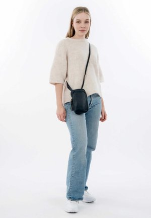 Borsa a tracolla nera con finitura testurizzata e dettaglio a nappina. Il modello indossa un maglione morbido di un beige chiaro e jeans blu, con sneakers bianche.