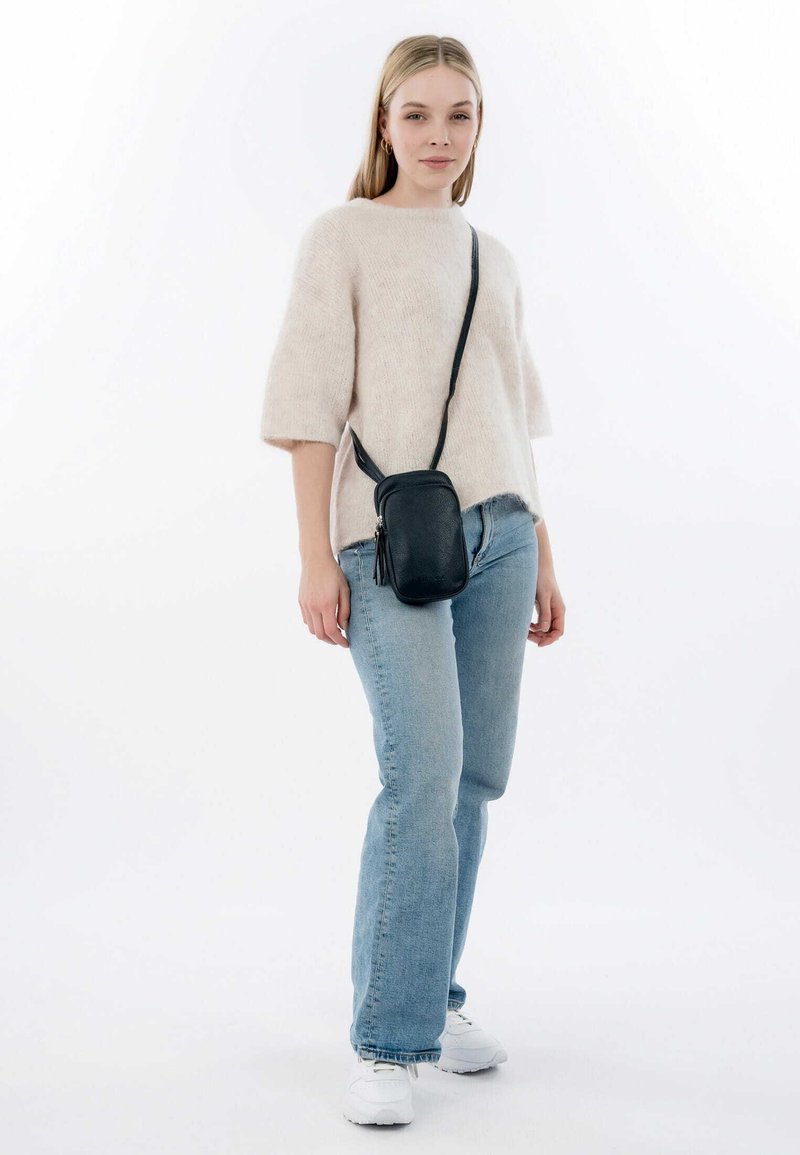 Black crossbody bag with a textured finish and tassel detail. Model wears a light beige fuzzy sweater and blue jeans, with white sneakers.