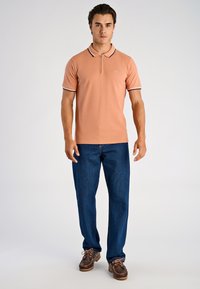 Jasnopomarańczowa koszulka polo z kołnierzem na zamek i czarno-białymi paskami, zestawiona z ciemnoniebieskimi dżinsami i brązowymi butami.