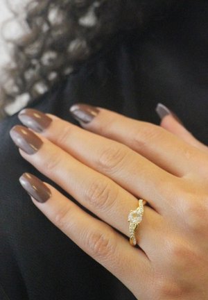 Bague en or avec une pierre transparente centrale, entourée d'un anneau torsadé orné de petits accents étincelants. Main reposant sur une surface sombre.