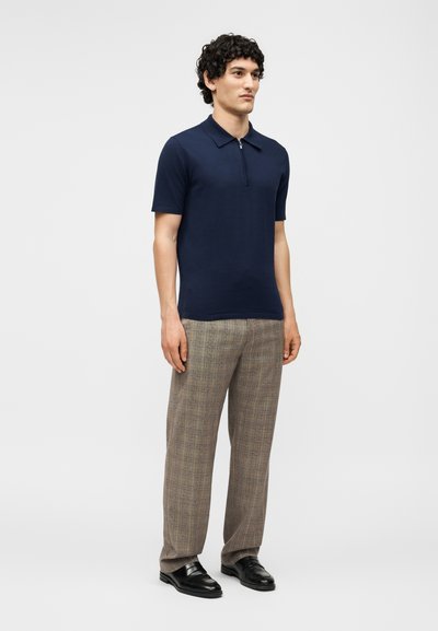 Άνδρας που στέκεται φορώντας ένα μπλε σκούρο πόλο με κοντά μανίκια και φερμουάρ στον γιακά, μπεζ καρό παντελόνια και μαύρα δερμάτινα μοκασίνια σε απλό φόντο.
