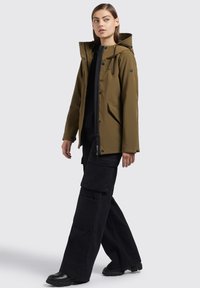 Giacca impermeabile verde oliva con cappuccio, chiusure a scatto frontali e tasche laterali. Indossata con pantaloni cargo neri a gamba larga e scarpe nere.