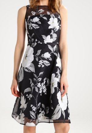 Robe noire sans manches avec motif floral blanc et gris, corsage ajusté et panneau transparent au niveau du décolleté, portée par une personne debout.