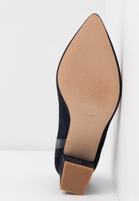 Chaussure à talon haut en daim bleu marine avec un bout pointu, un talon conique et une semelle en caoutchouc texturée. Dispose d'une bande d'accent noire lisse.
