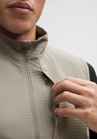 Beige prošívaná vesta s vysokým límcem, s předním zipem. Jedna ruka táhne za zip, viditelný detail vzoru látky.