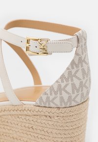 Gros plan sur une sandale compensée beige avec bride à la cheville, boucle dorée MK, plateforme tissée et motif logo MK sur la partie talon.