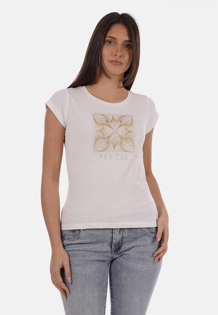 Jeune femme portant un t-shirt blanc avec un motif géométrique doré et le texte "YES ZEE", associé à un jean denim gris clair, debout les mains détendues.