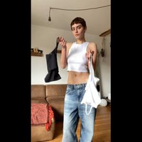 Duas camisolas são exibidas: uma preta e uma branca. O modelo usa um top curto branco e jeans largos de tom azul claro. O tecido apresenta uma textura visível.
