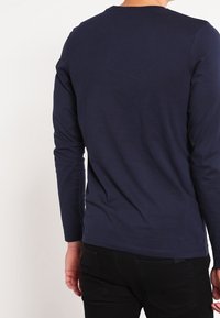 Tmavě modré tričko s dlouhým rukávem vyrobené z měkké bavlny, s kulatým výstřihem a přiléhavým střihem, zobrazené ze zadní strany. Viditelné černé džíny.