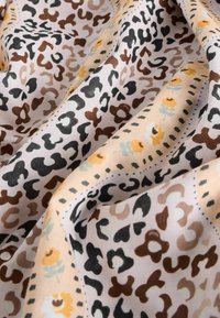 Tkanina s uzorkom leoparda u bež boji, koja sadrži crne i smeđe mrlje, s cvjetnim akcentima u žutoj i svijetloplavoj boji duž detaljno ukrašenog ruba.