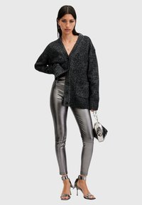 Cardigan nero a maglia spessa con scollatura a V e chiusura a bottone singolo; abbinato a leggings metallici argento e tacchi a spirale argentati.