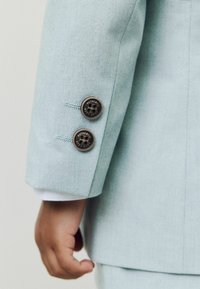 Rokav svetlo modrega blazerja z dvema rjavima gumboma z napisom »AUTHENTIC NEXT DUDE«, ki ga nosi oseba z vidno roko in manšetom bele srajce.