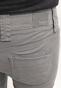 Gros plan sur un pantalon gris Pepe Jeans montrant la poche arrière, la passants de ceinture et l’étiquette en tissu cousue au-dessus de la poche.