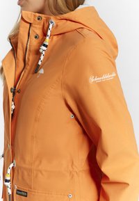 Veste imperméable orange avec capuche à cordon de serrage, accents argentés, logo sur la manche et devant zippé. Dotée d'un tissu texturé.