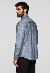 Homme aux cheveux foncés et à la barbe portant une chemise à manches longues grise à motif floral, tourné de profil, faisant face à gauche sur un fond uni.