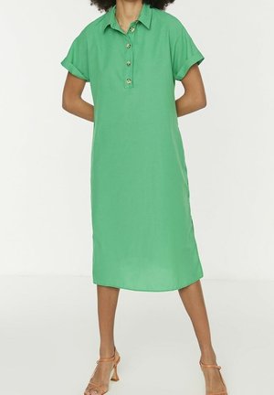 Femme portant une robe midi verte à manches courtes, boutonnée avec col, associée à des sandales à talons hauts orange, debout les mains dans le dos.