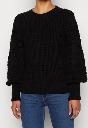 Femme portant un pull en maille noire avec des manches bouffantes texturées et semi-transparentes et un jean bleu, montrée des épaules aux hanches.