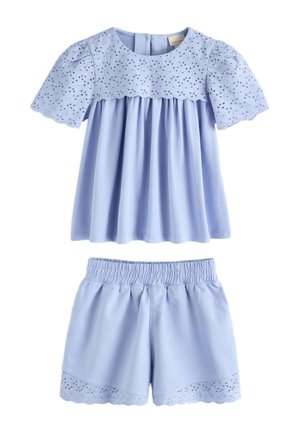 Junges Mädchen mit langen dunklen Haaren, das ein hellblaues Spitzen-Top und passende Shorts trägt, steht vor einem einfarbigen beigen Hintergrund.