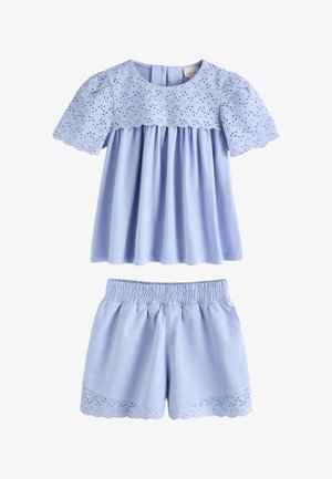 Jeune fille aux longs cheveux foncés portant un haut en dentelle bleue claire avec des œillets et un short assorti, debout devant un fond beige uni.