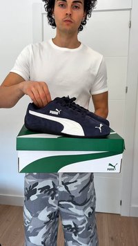 Homem com camisa branca e calças de camuflagem segura uma caixa de sapatos verde da Puma com ténis da Puma em azul marinho e branco em cima.