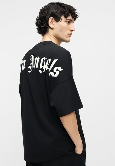 Bărbat tânăr cu păr scurt și creț, purtând un tricou negru larg cu litere gotice albe pe spate, stând în fața unui fundal simplu.