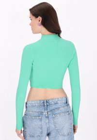 Minzgrünes, geripptes, langärmliges Crop-Top mit Stehkragen; kombiniert mit hochgeschnittenen, hellblauen Jeans mit Gesäßtaschen.