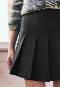 Personne portant une jupe noire plissée et un pull en maille vert à motifs avec des manches retroussées, se tenant à l'intérieur près d'une fenêtre.