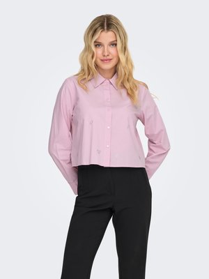 Femme blonde aux cheveux ondulés portant une chemise rose clair à manches longues boutonnée et un pantalon noir, debout les mains dans le dos.