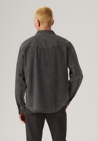 Camisa de mezclilla negra con cuello, mangas largas y detalles de costura visibles. Presenta un corte recto y un dobladillo ligeramente curvado en la parte posterior.