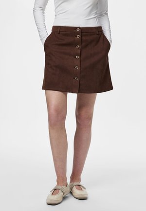 Personne portant une jupe marron à boutons sur le devant avec des poches, un haut blanc à manches longues et des chaussures style Mary Jane beige, debout les mains dans les poches.