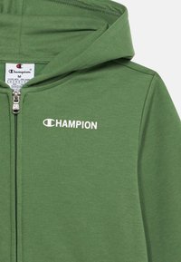 Vihreä vetskarilla varustettu huppari pehmeästä kankaasta, jossa on huppu. Rinnassa on valkoinen "CHAMPION"-logo ja vetoketjukiinnitys.