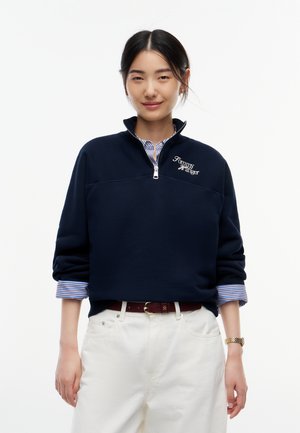 Ung kvinde iført en mørkeblå Tommy Hilfiger sweatshirt med kvart-lynlås, stribede skjorteærmer der stikker frem, hvide bukser og et guldarmbåndsur.
