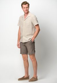 Camicia a maniche corte in lino beige chiaro con una tasca sul petto, abbinata a pantaloni corti marroni con texture e scarpe slip-on beige.