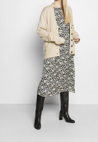 Cárdigan de punto beige con botones, llevado sobre un vestido negro de estampado floral. Botas de tacón hasta la rodilla en negro completan el conjunto.