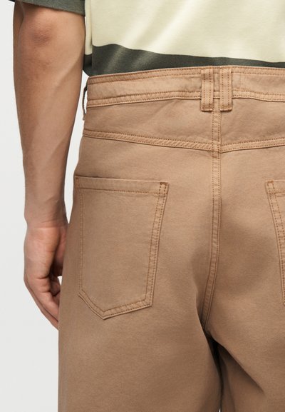 Κοντινή εικόνα στο κάτω μέρος της πλάτης ενός ατόμου που φοράει καφέ παντελόνι με τσέπες και ριγωτό πράσινο, λευκό και μπεζ πουκάμισο.