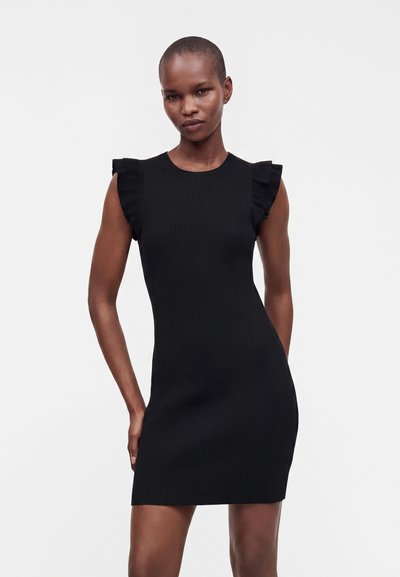 Femme aux cheveux courts portant une robe noire côtelée sans manches avec des volants aux épaules, debout devant un fond blanc uni.