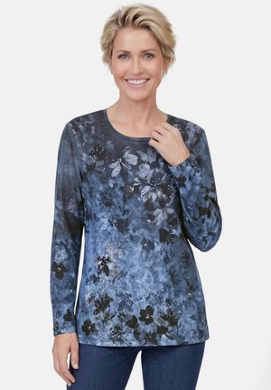 Femme souriante aux cheveux blonds courts portant une chemise bleue à motif floral à manches longues et un jean bleu sur un fond uni.